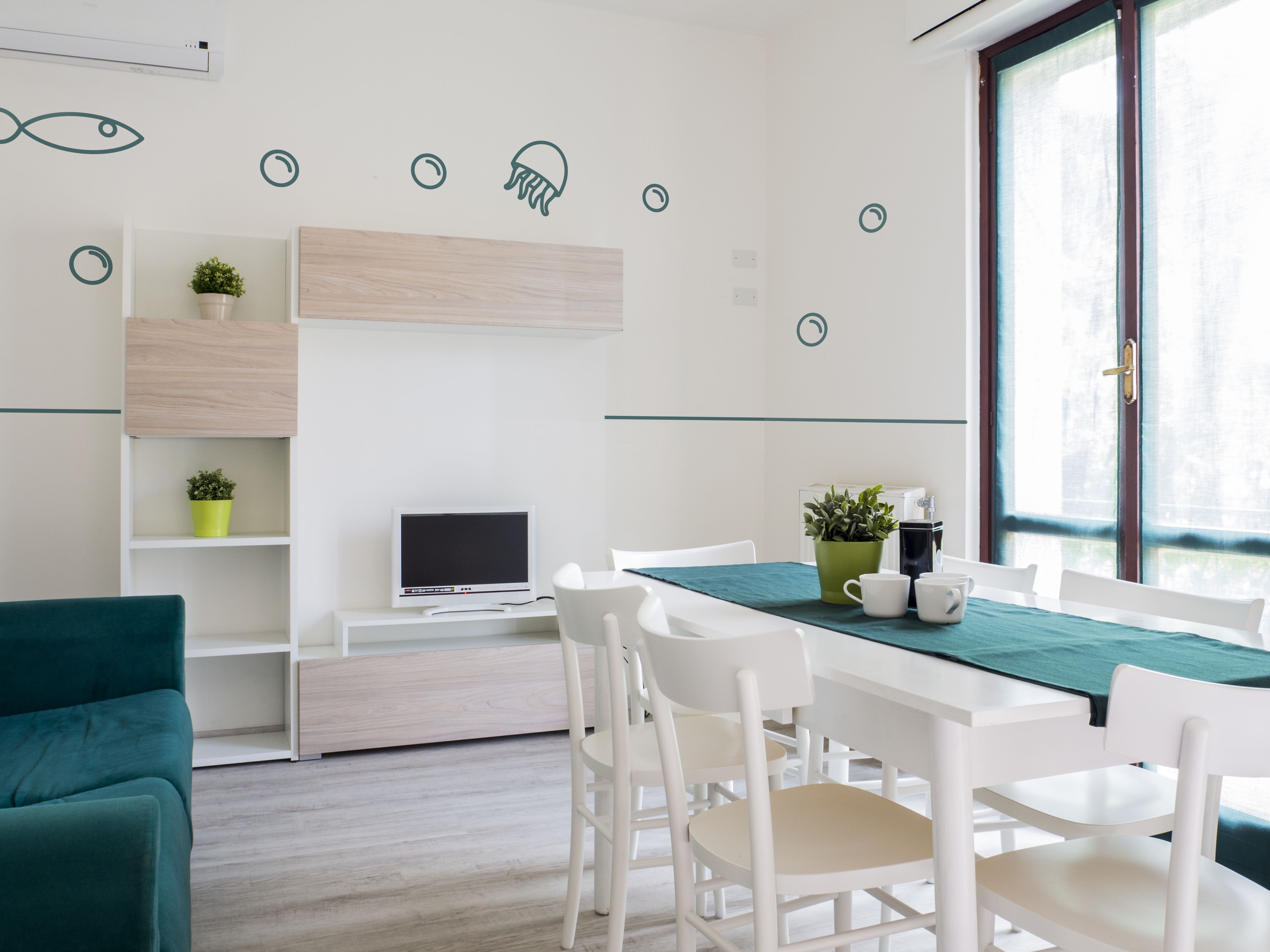 Une chambre moderne avec un bureau blanc, un téléviseur à écran plat, des chaises blanches, une nappe turquoise et une fenêtre donnant sur un paysage agréable.
