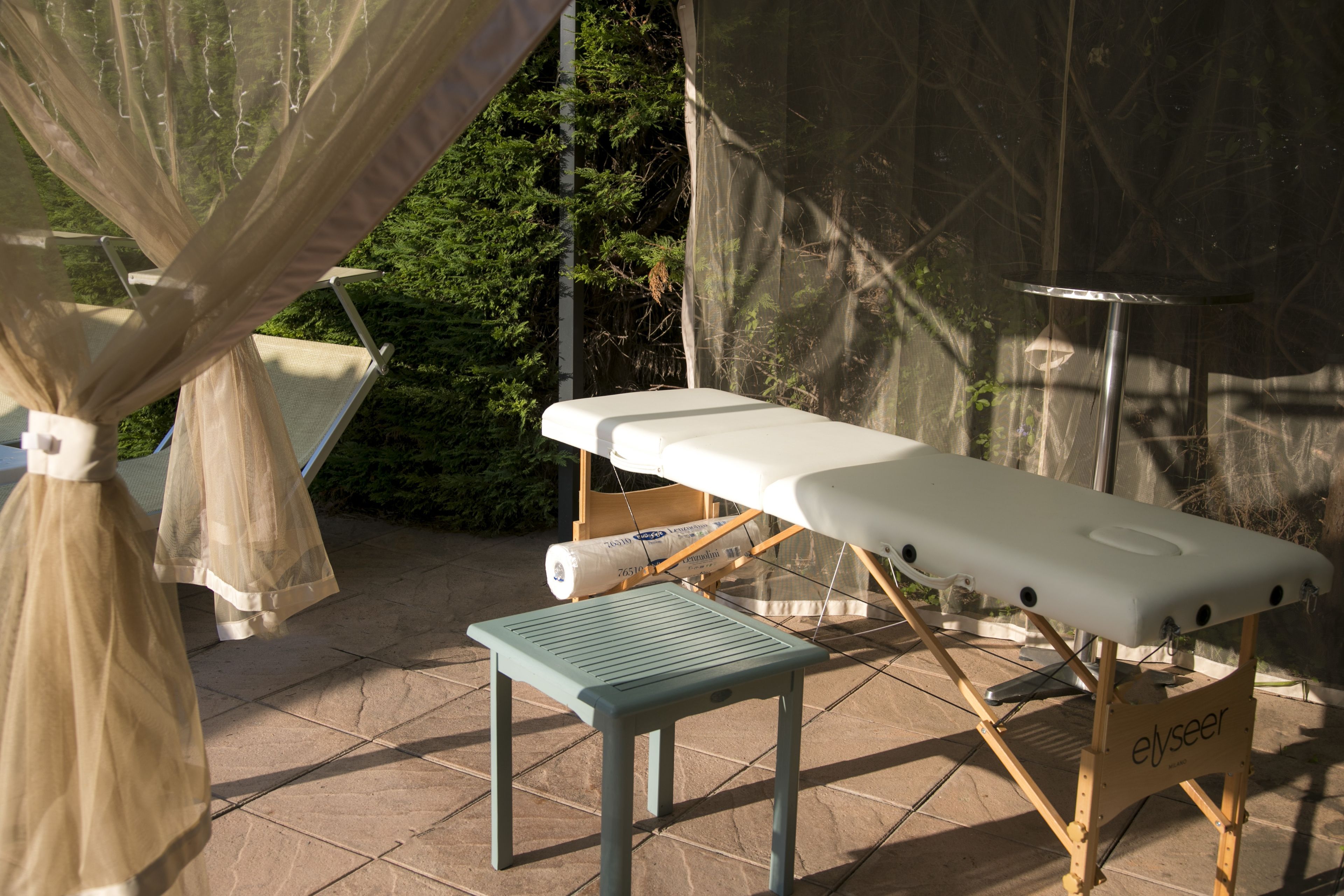 A massage table and a small stool in an outdoor setting with a tiled floor and a canopy.
