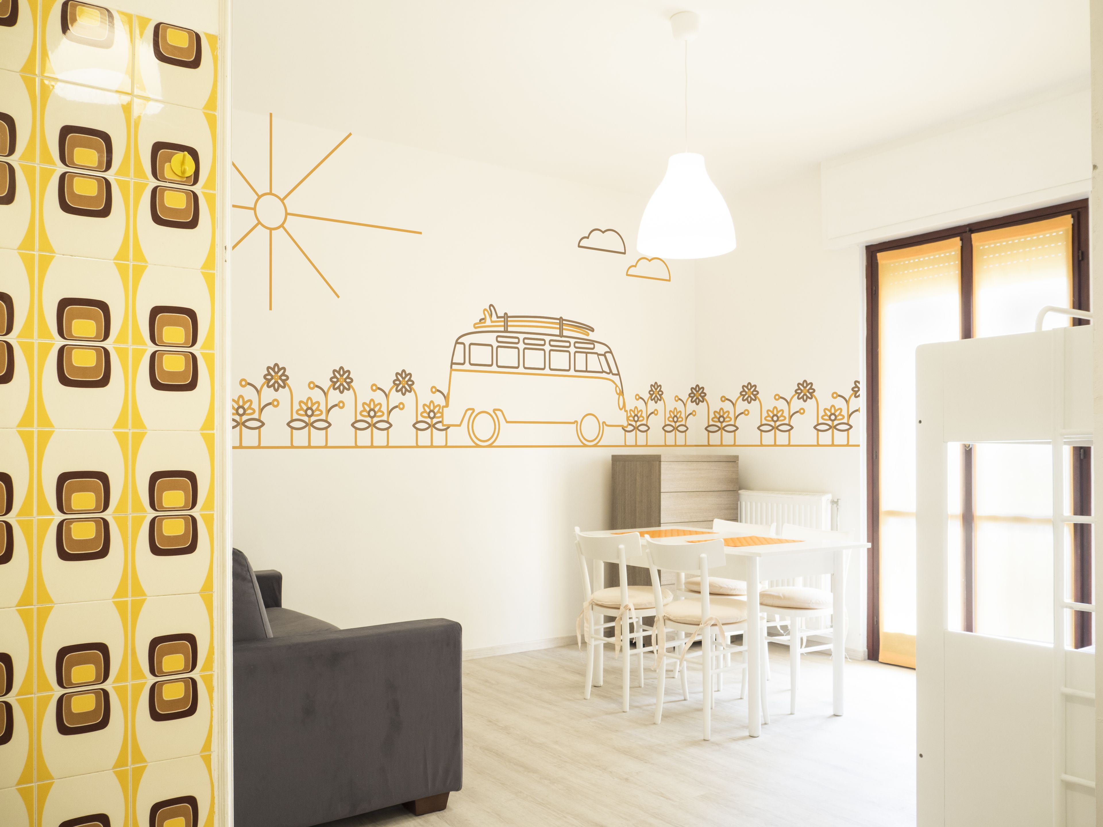 Una sala moderna con un sofá, una mesa de comedor con sillas, y una pared con un diseño de sol, nubes, flores y un autobús.