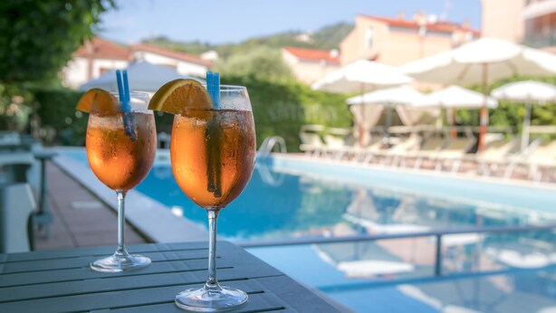 To glas med drikkevarer, et appelsinstykke og et sugerør, placeret på et bord ved en pool med parasoller og liggestole i baggrunden.