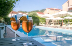 Deux verres contenant une boisson, décorés d’une tranche d’orange et munis d’une paille, sont posés sur une table au bord d’une piscine, avec des parasols et des chaises longues en arrière-plan.