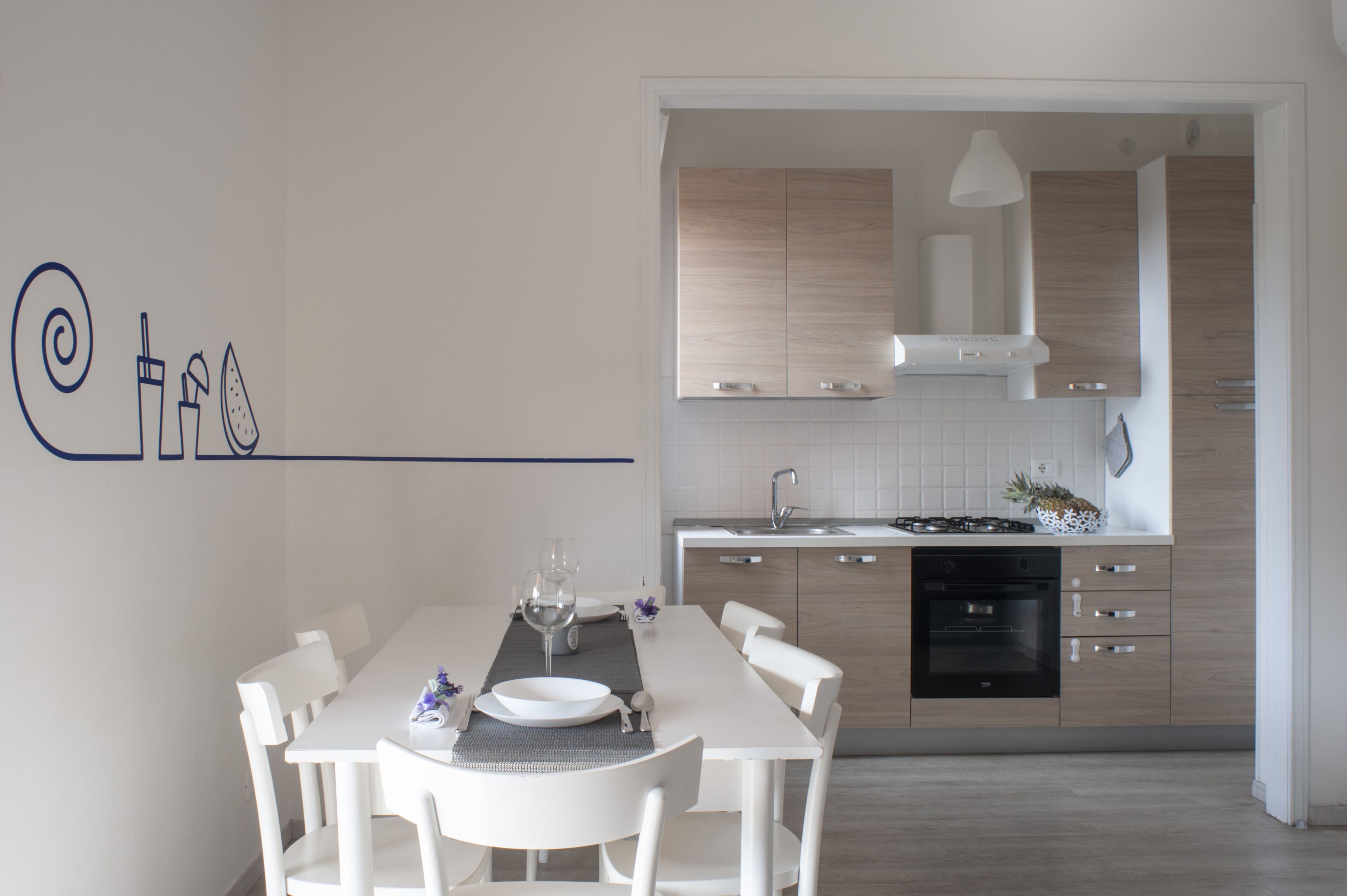 Cocina moderna con armarios blancos, horno empotrado y zona de comedor con mesa y sillas blancas.