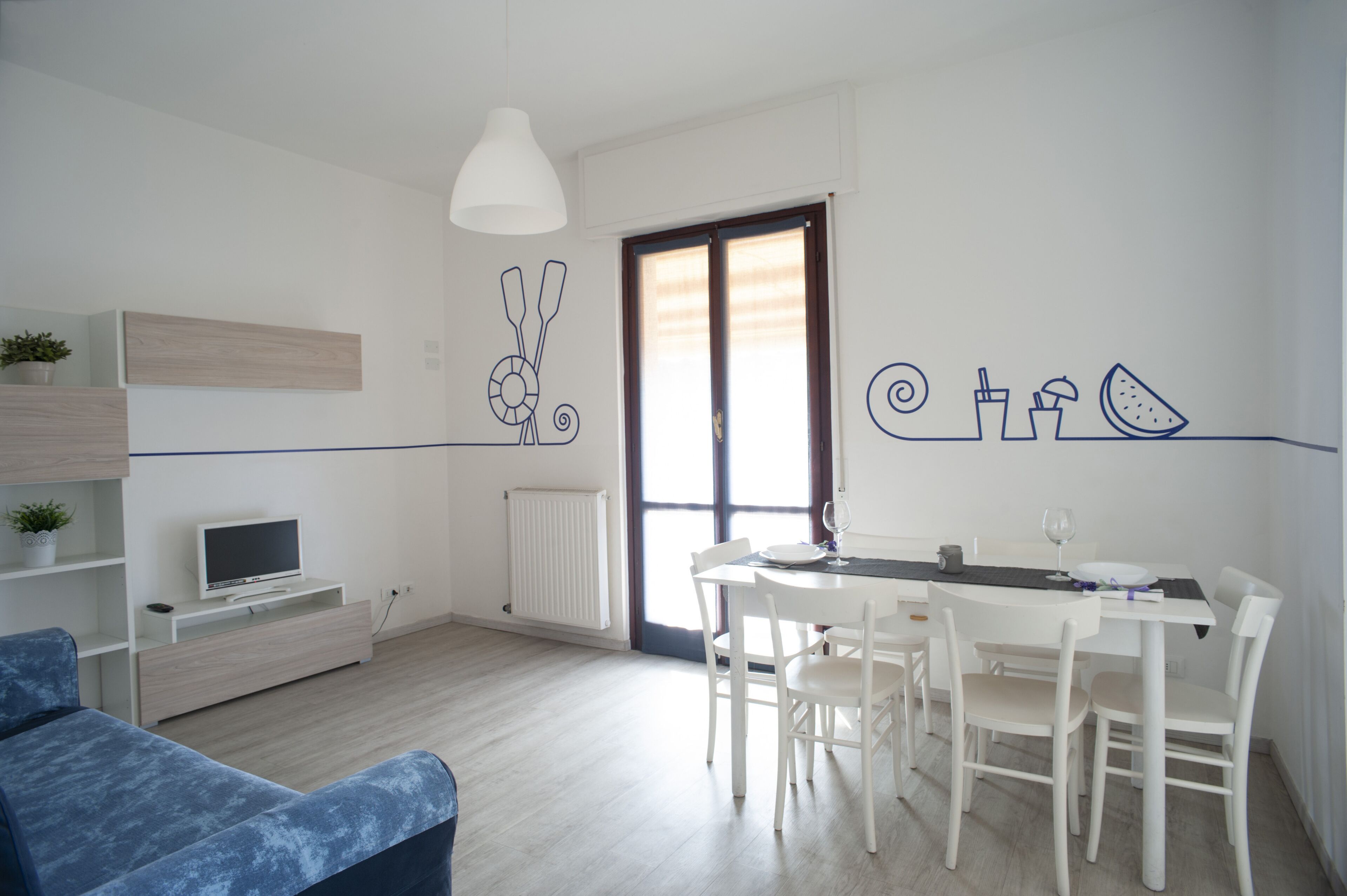 A modern living room with a blue sofa, a dining table with white chairs, a white TV on a stand, and a large window with blinds.