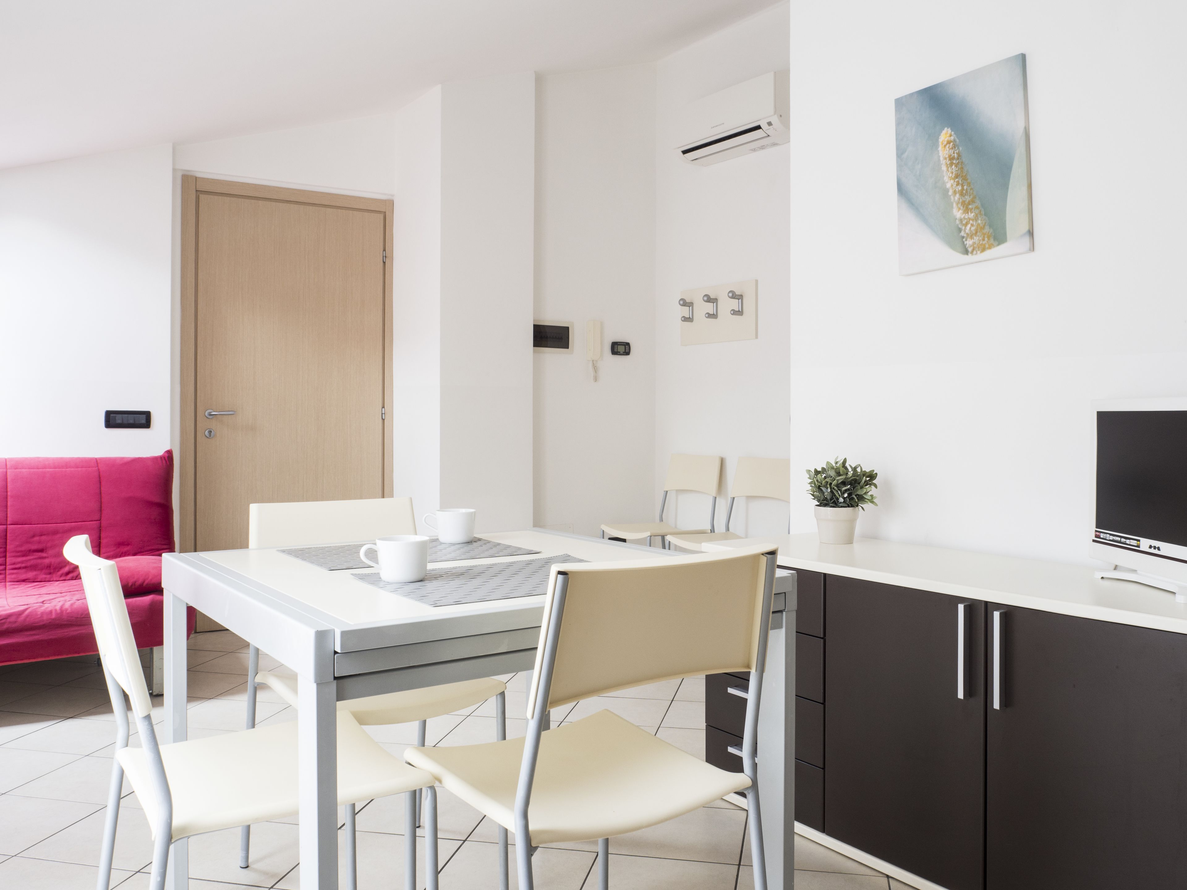 Quarto moderno e bem iluminado, com mesa de jantar, cadeiras, sofá, televisão e um vaso de planta.