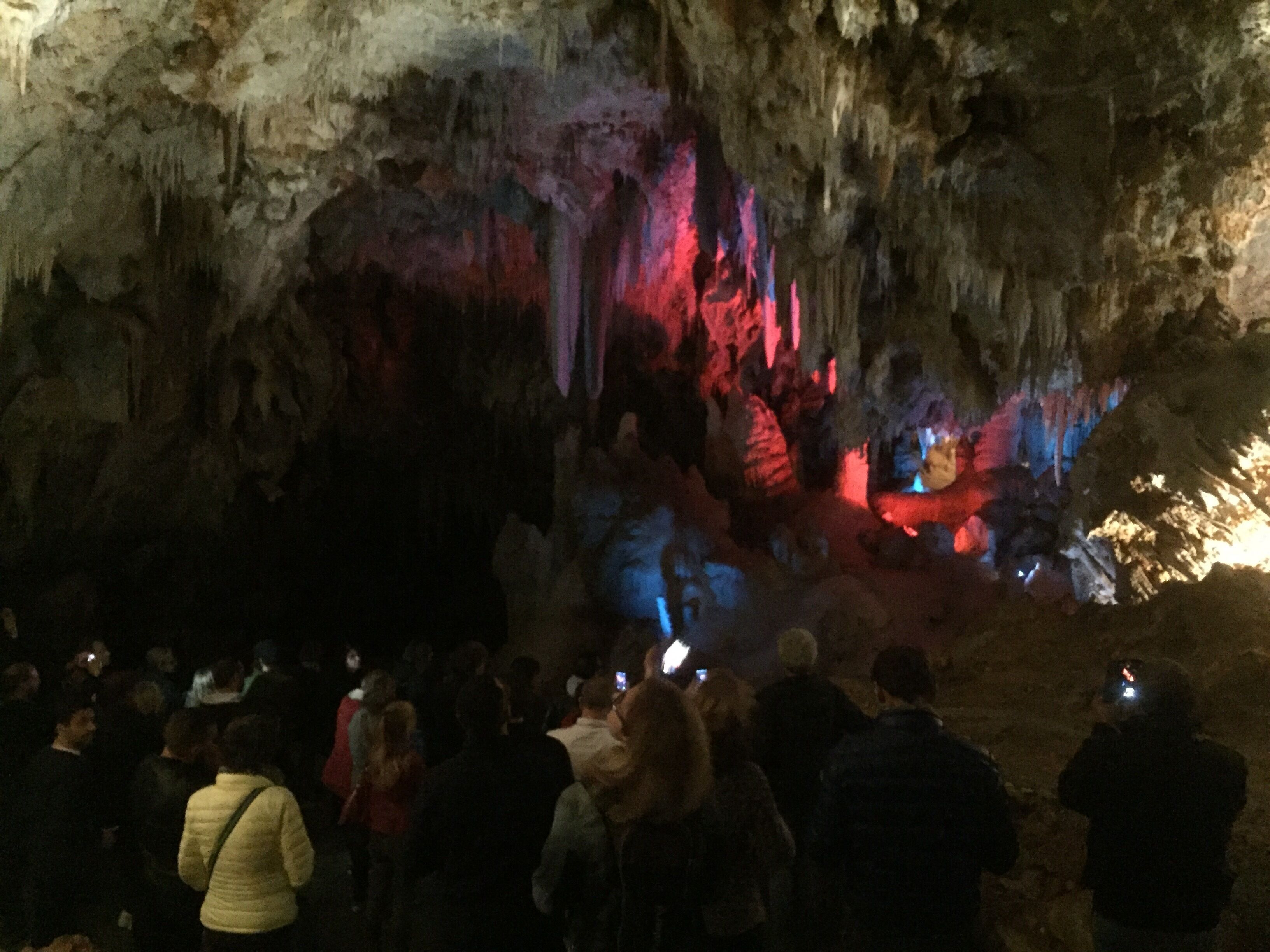 A cave interior with stalactites and stalagmites, illuminated by artificial lighting.