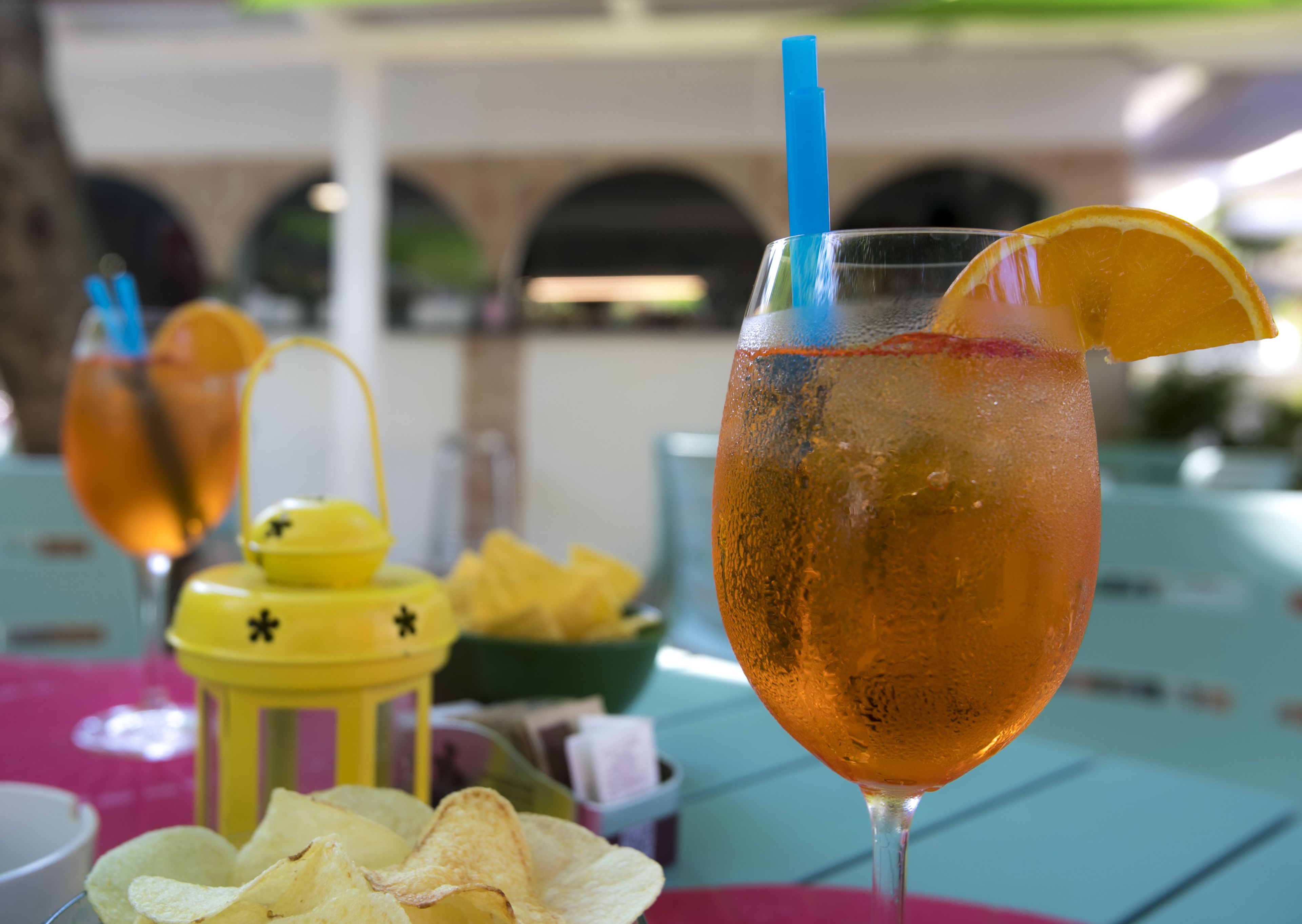 A table with a glass of iced drink with an orange slice and a blue straw, surrounded by yellow items and chips.