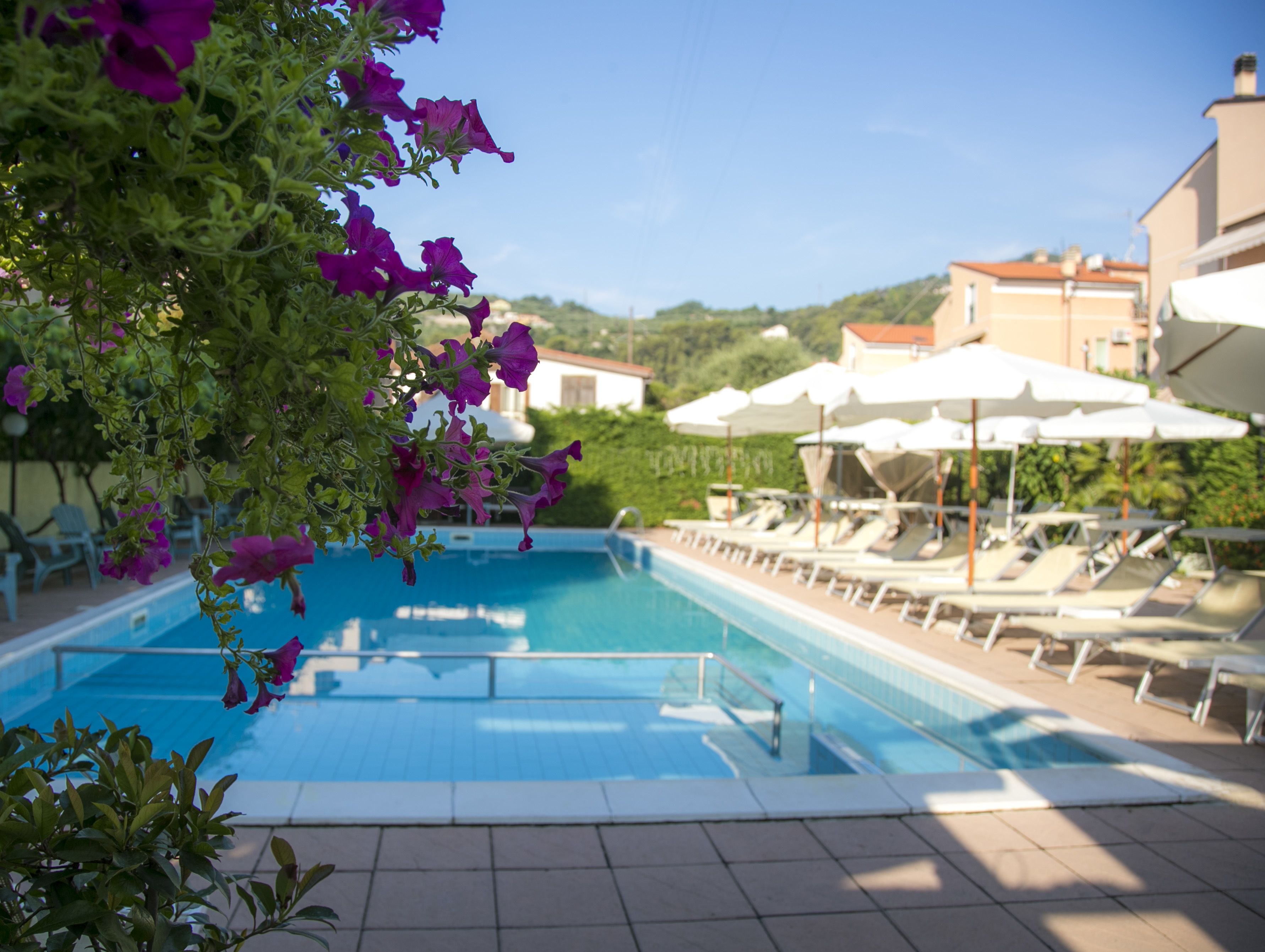 Ein Schwimmbecken mit Liegestühlen und Sonnenschirmen, umgeben von Grünflächen und Gebäuden.