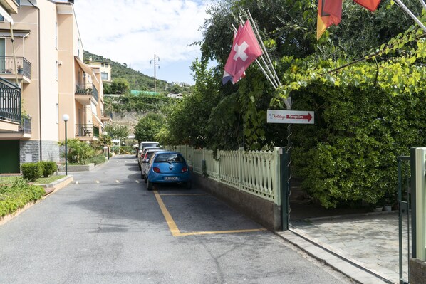 Une rue bordée de voitures garées, une enseigne indiquant un lieu d’accueil pour les vacances, et des drapeaux.
