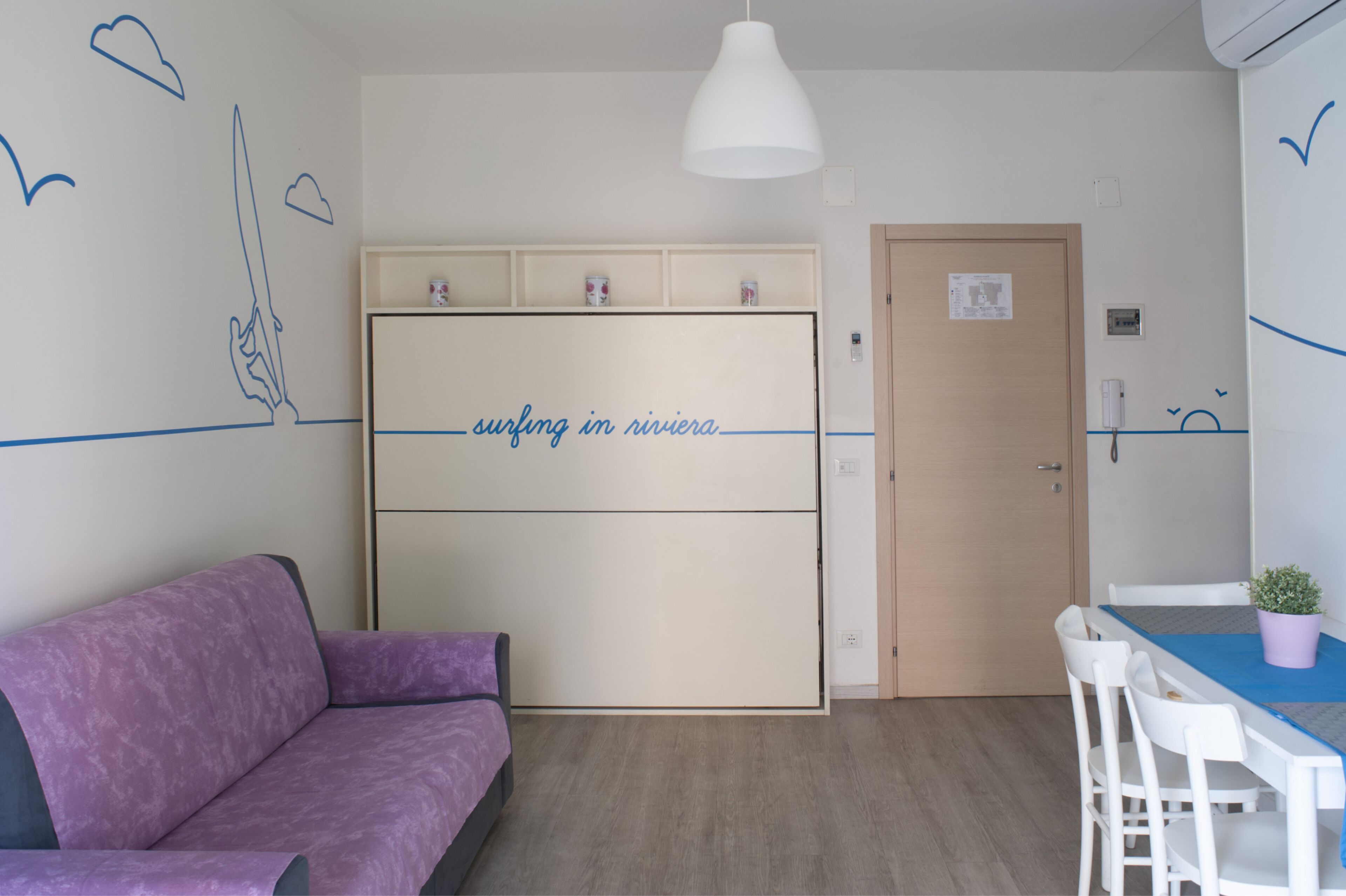Ein Zimmer mit einem violetten Sofa, einem weißen Tisch mit Stühlen, einer weißen Pendelleuchte und einem Schrank mit dem Aufdruck 'Surfen in Riviera'.