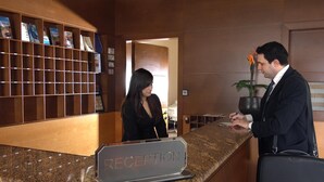 A reception desk with a 'RECEPTION' sign, a woman standing behind it, and a man holding a pen and a notepad. - Byzantio Hotel (Pavlos Melas)