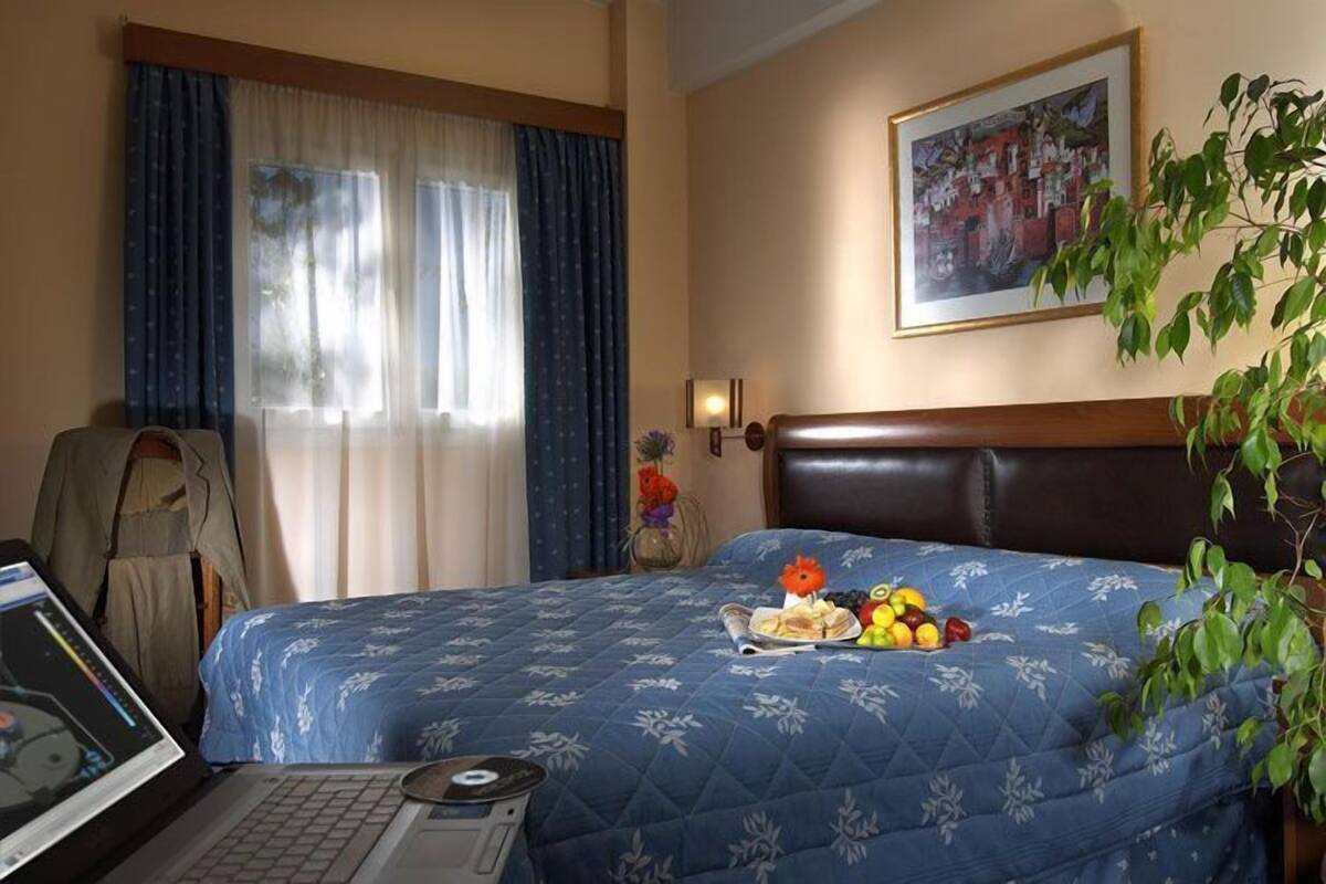 A hotel room with a bed, a laptop, a plant, and a painting on the wall.