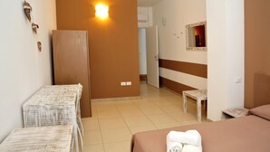 A hotel room with a bed, a small table, a chair, a wall-mounted light, and a door leading to another room. - La Punta (Otranto)
