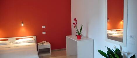 Una habitación de hotel con una pared de acento roja, una cama con ropa de cama blanca, una mesita de noche, una pequeña mesa con una maceta roja y una planta, y un espejo en la pared.