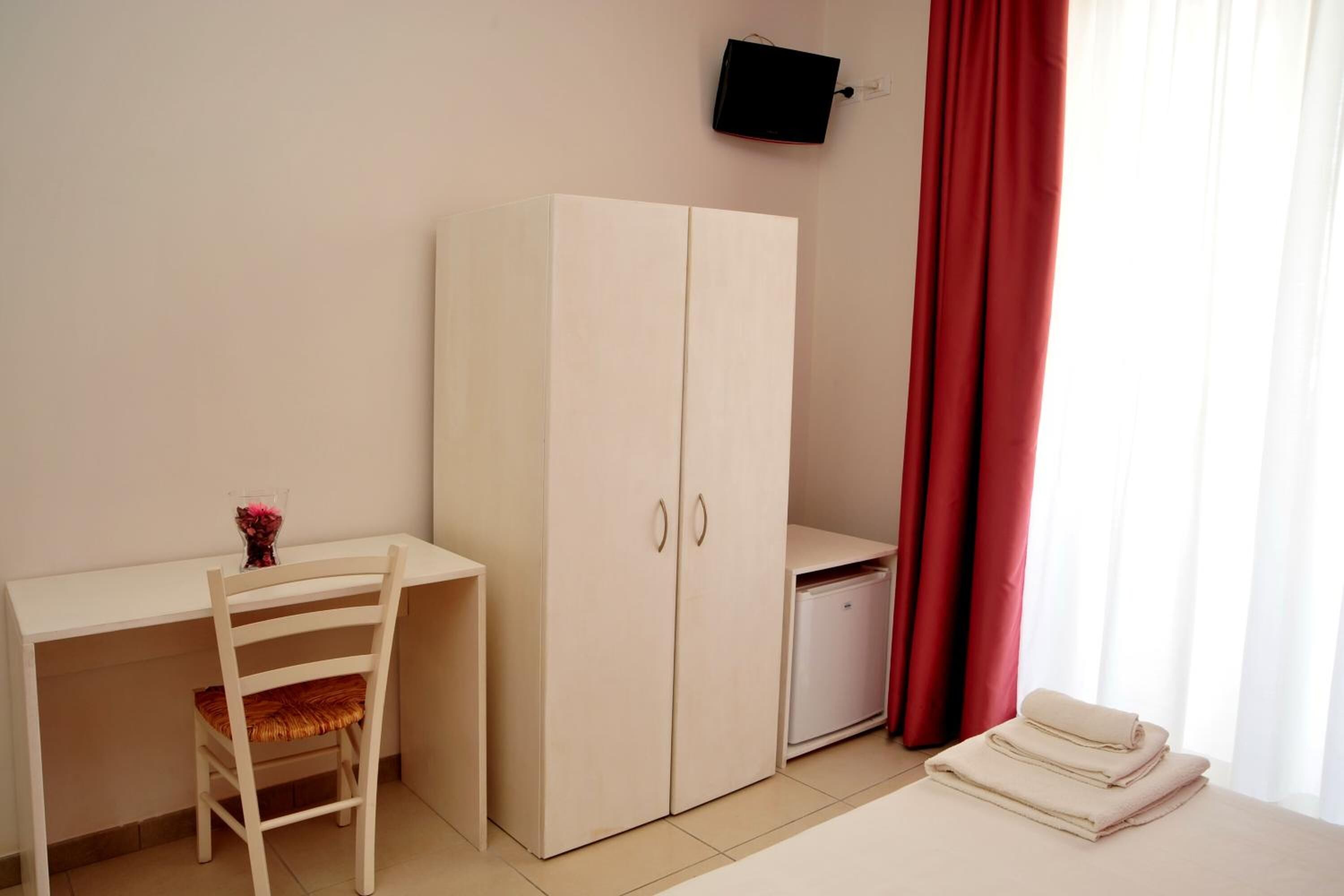 Ein Zimmer mit Schreibtisch, Stuhl, Kleiderschrank, Fernseher und einem fenster mit Vorhängen.