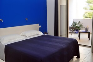 A hotel room with a bed, a nightstand, a potted plant, and a view of the outdoors.