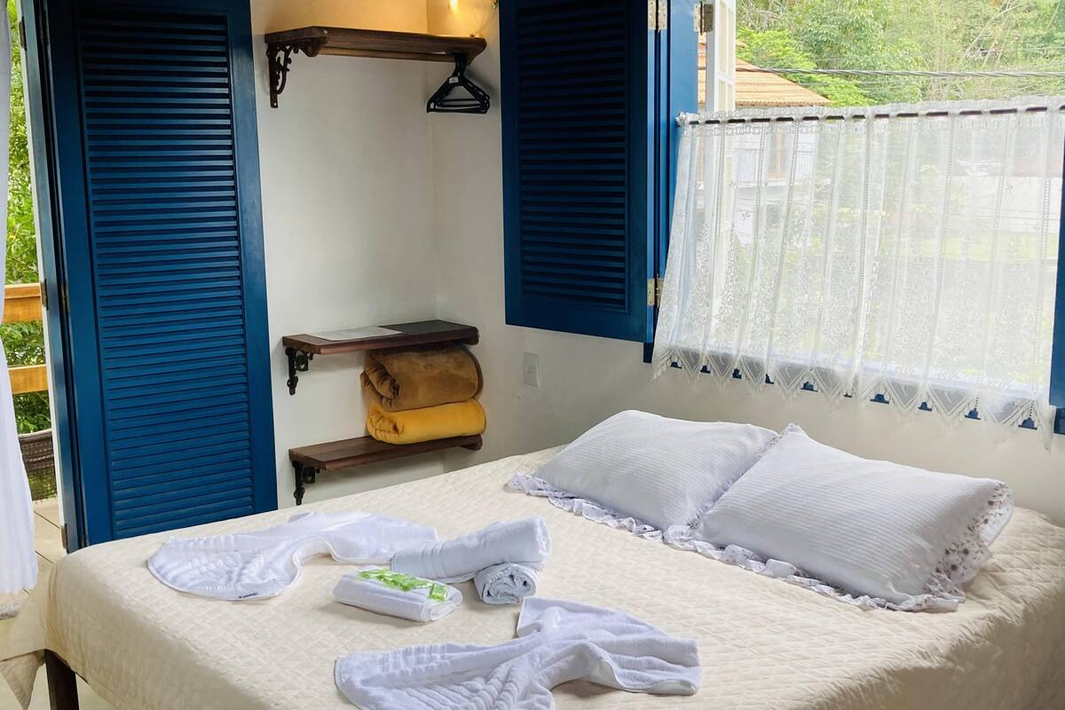 A neatly made bed with white linens and pillows, a wooden shelf with a rolled-up blanket, and a window with lace curtains.