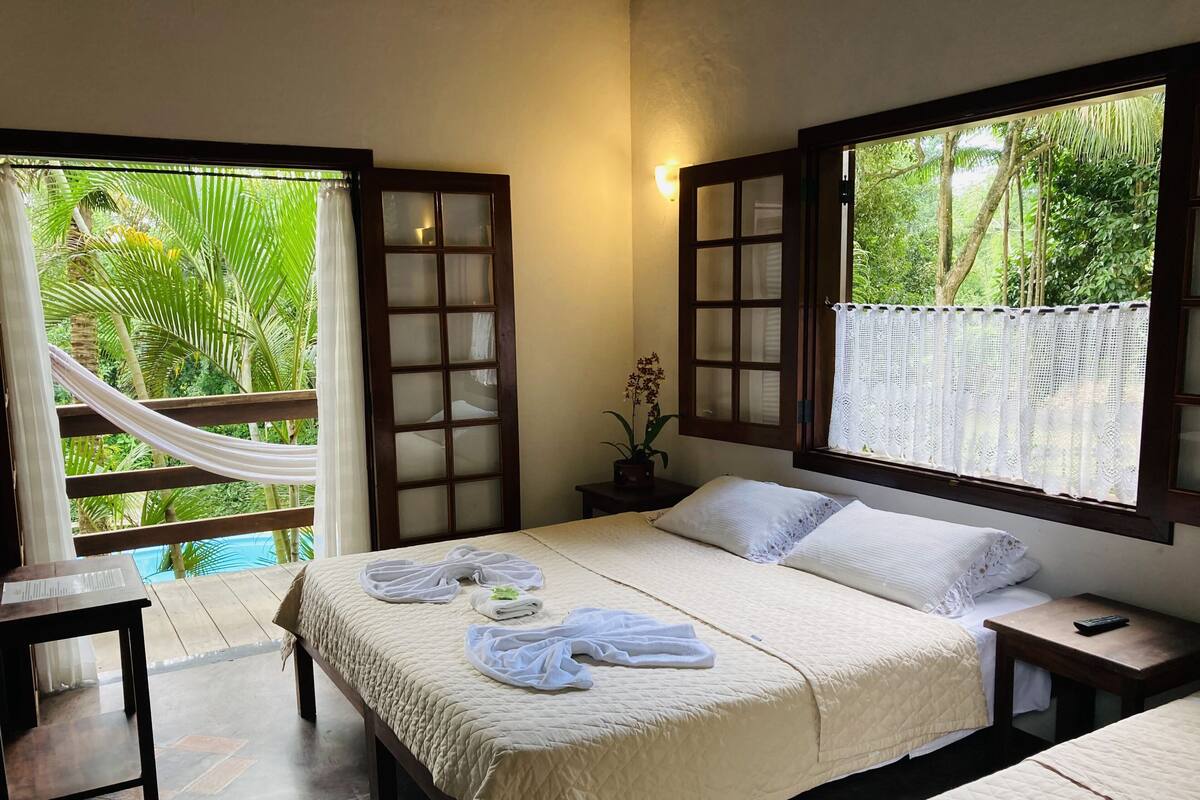 A bedroom with a bed, a nightstand, a small table, a window with a view of greenery, and a hammock.