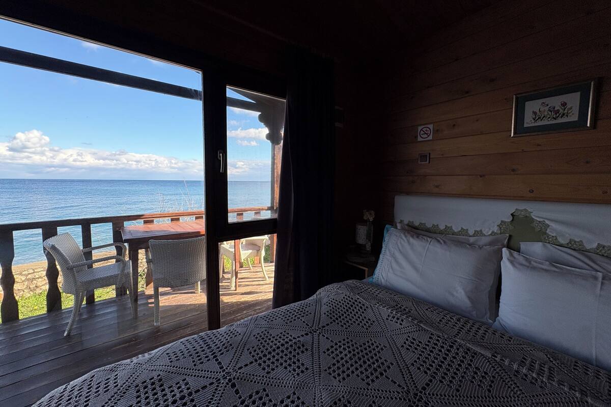 A bedroom with a large window overlooking a beach, a wooden deck with chairs, and a bed with a patterned blanket.