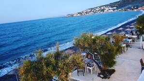 A coastal view with a beach, a mountain in the background, and a row of outdoor seating with umbrellas. - Lipsos Hotel Ata's Place (Karaburun)
