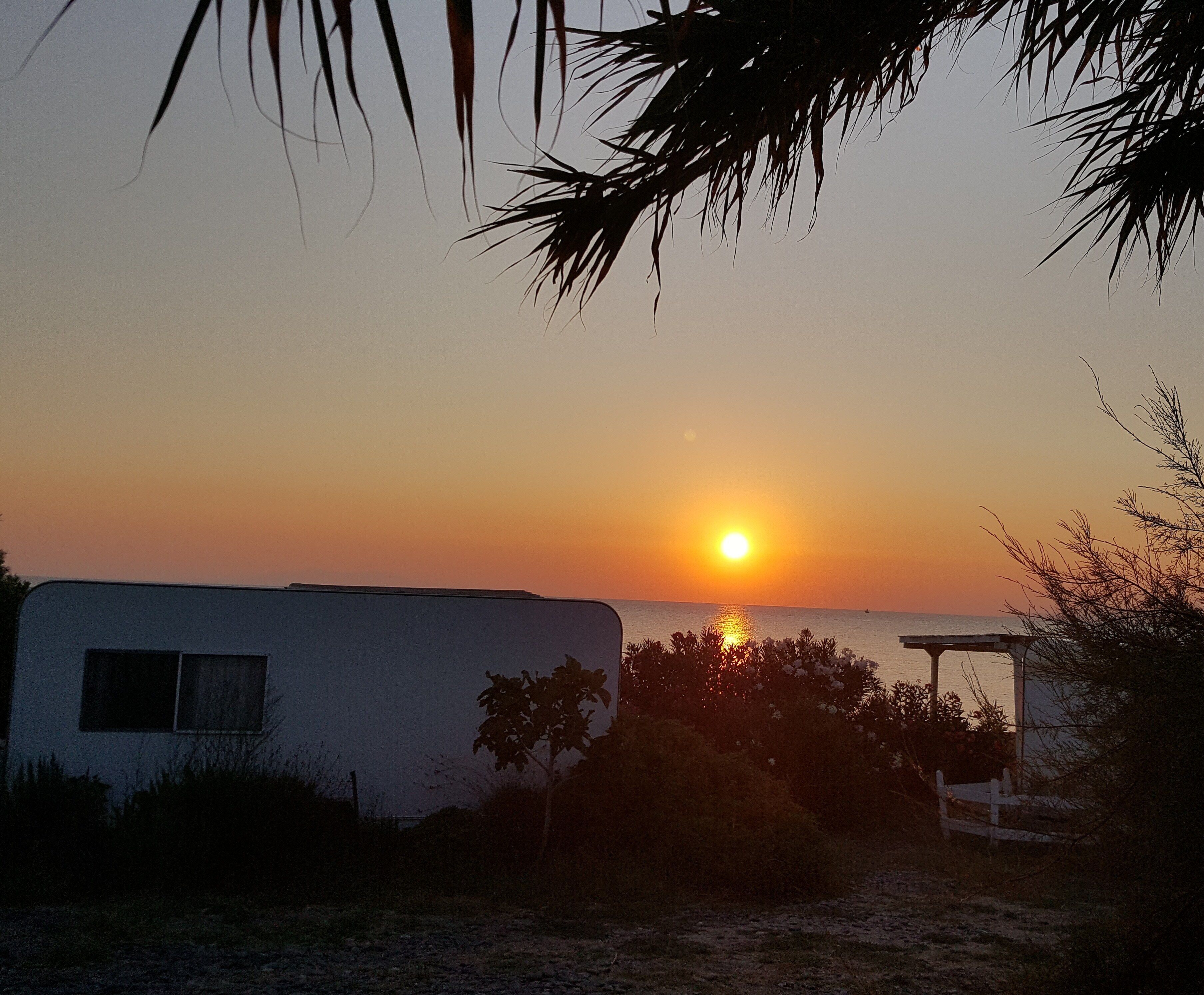 Ein Sonnenuntergang über einem Gewässer, im Vordergrund ein Wohnwagen und eine weiße Umzäunung.