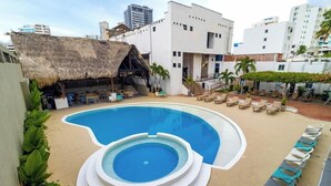 A swimming pool with a unique shape, surrounded by lounge chairs and a bar area with a thatched roof. - Sansiraka (Santa Marta)