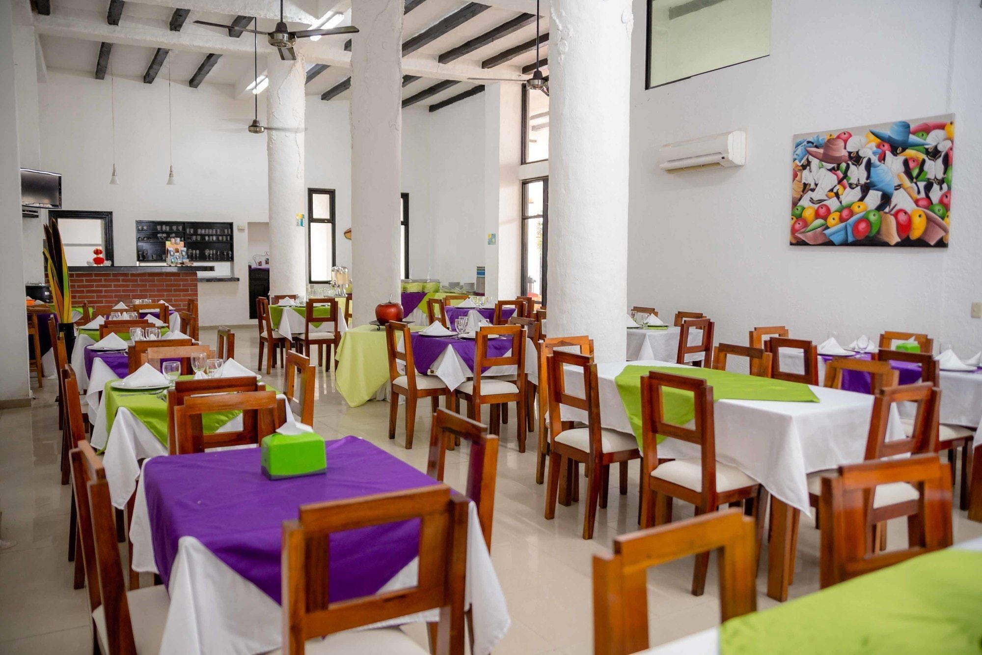 A dining area with multiple tables and chairs, some with tablecloths. There are ceiling fans and a painting on the wall.