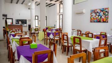 A dining area with multiple tables and chairs, some with tablecloths. There are ceiling fans and a painting on the wall.