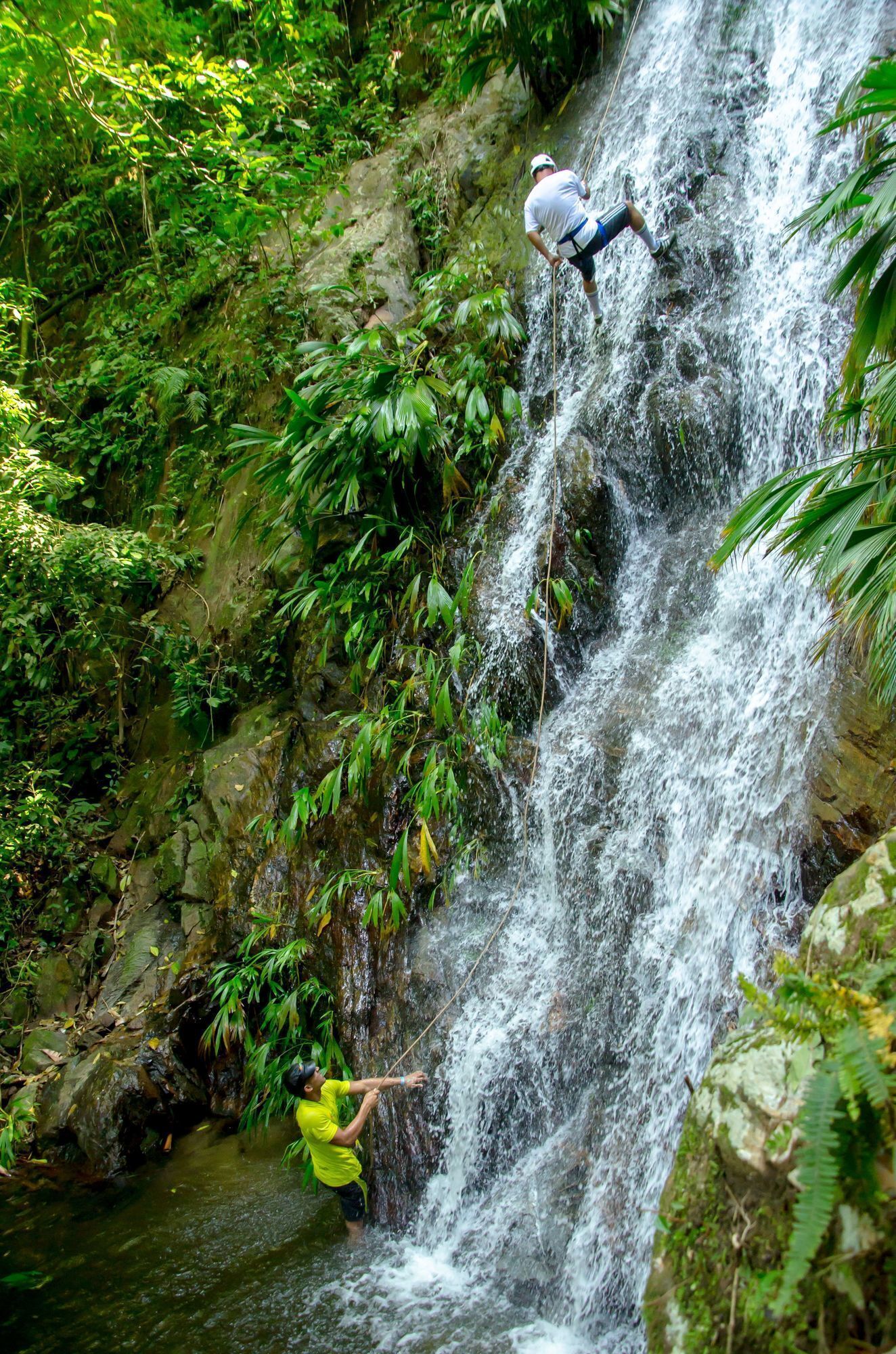 Een waterval met weelderige begroeiing en twee mensen die eraan klimmen.