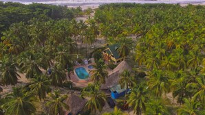 An aerial view of a tropical resort with a swimming pool, thatched-roof structures, and dense palm trees. - Sansiraka (Santa Marta)