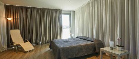 Een moderne hotelkamer met een groot bed, een zithoek met een stoel, een kleine tafel en een raam met gordijnen.