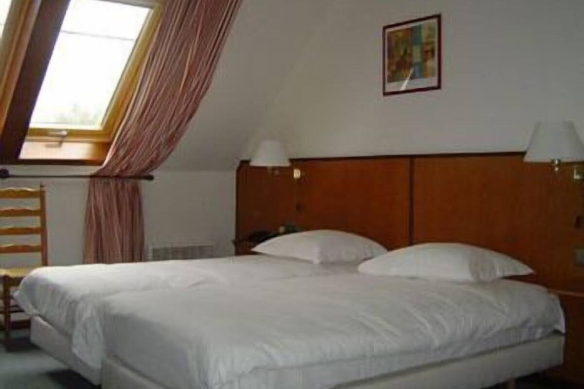 A hotel room with a large bed, two bedside lamps, a window with curtains, and a painting on the wall.