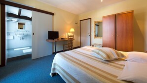 A hotel room with a bed, a desk, a chair, a television, a wardrobe, and a bathroom visible through an open door. - Auberge le XIXème (Hensies)