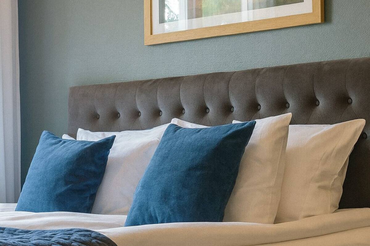 Cama bem arrumada com cabeceira estofada, quadro na parede e manta decorativa.