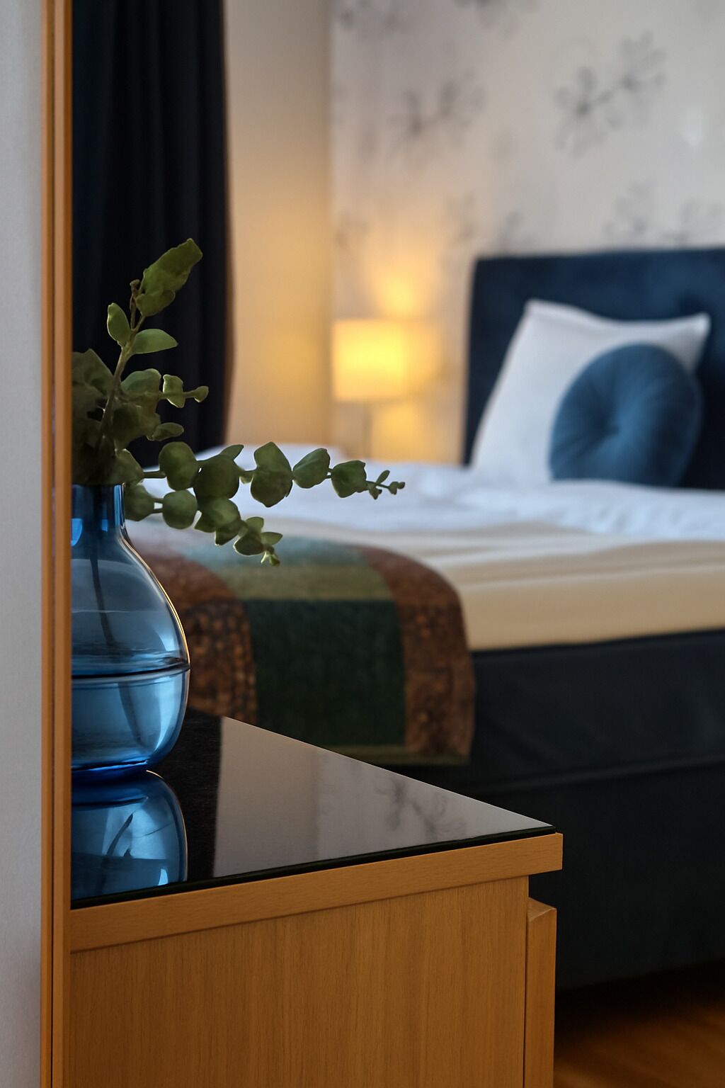 Una camera d'albergo con un comodino in legno, un vaso blu con verdeggianti piante, un letto con testiera scura, cuscini bianchi e una coperta verde.