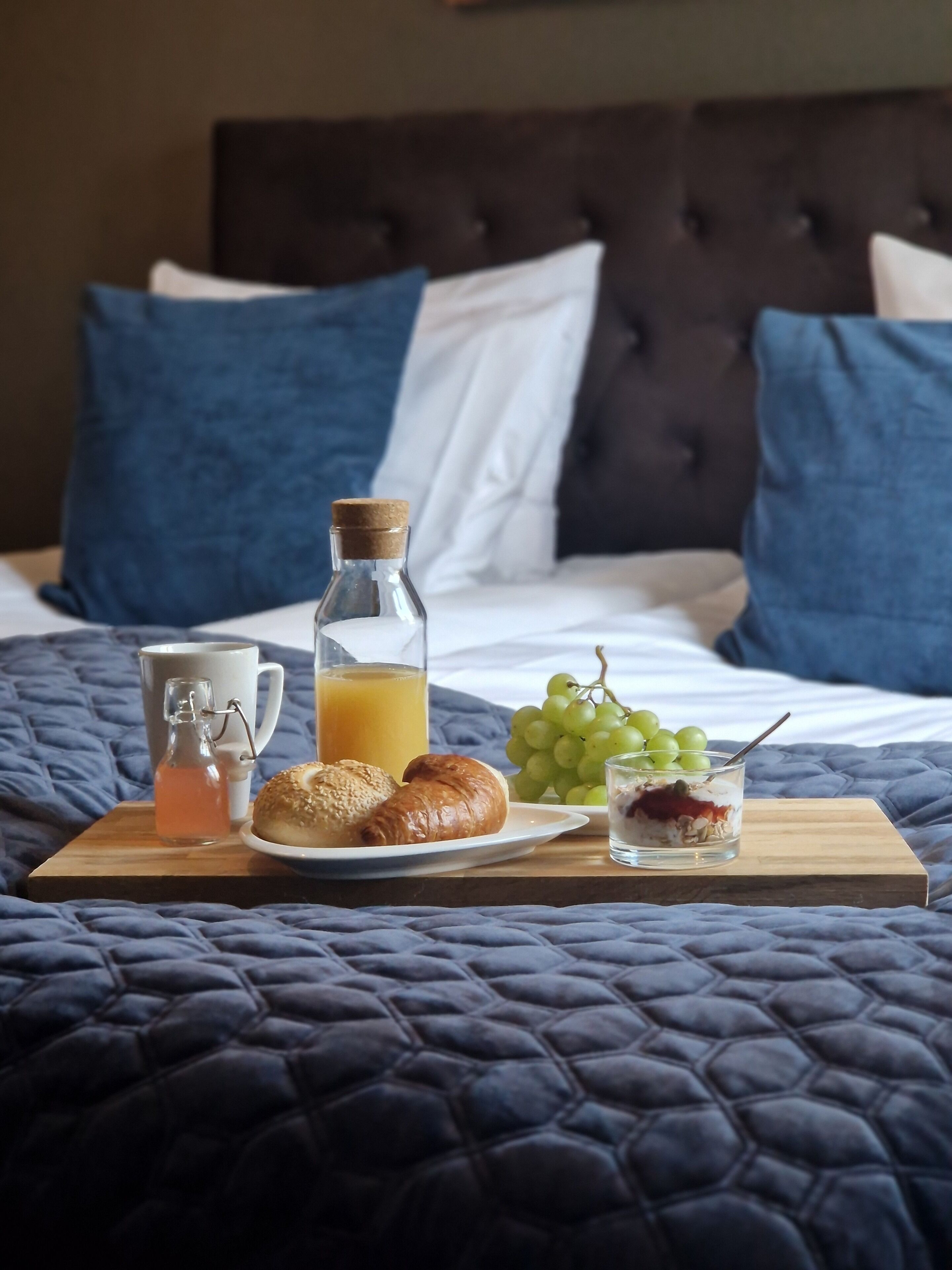 A neatly made breakfast tray on a bed with a dark blue quilt, including croissants, grapes, a glass of orange juice, and a small jar of jam.