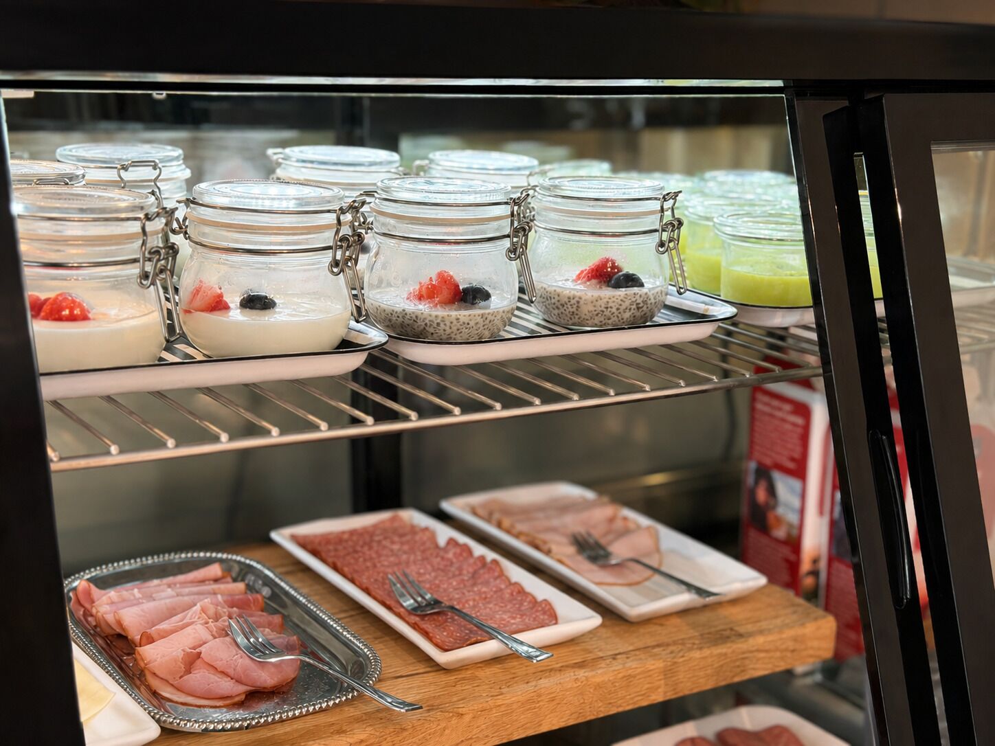 A refrigerated display case with various food items, including jars of yogurt with fruits, chia seed pudding, and sliced meats.