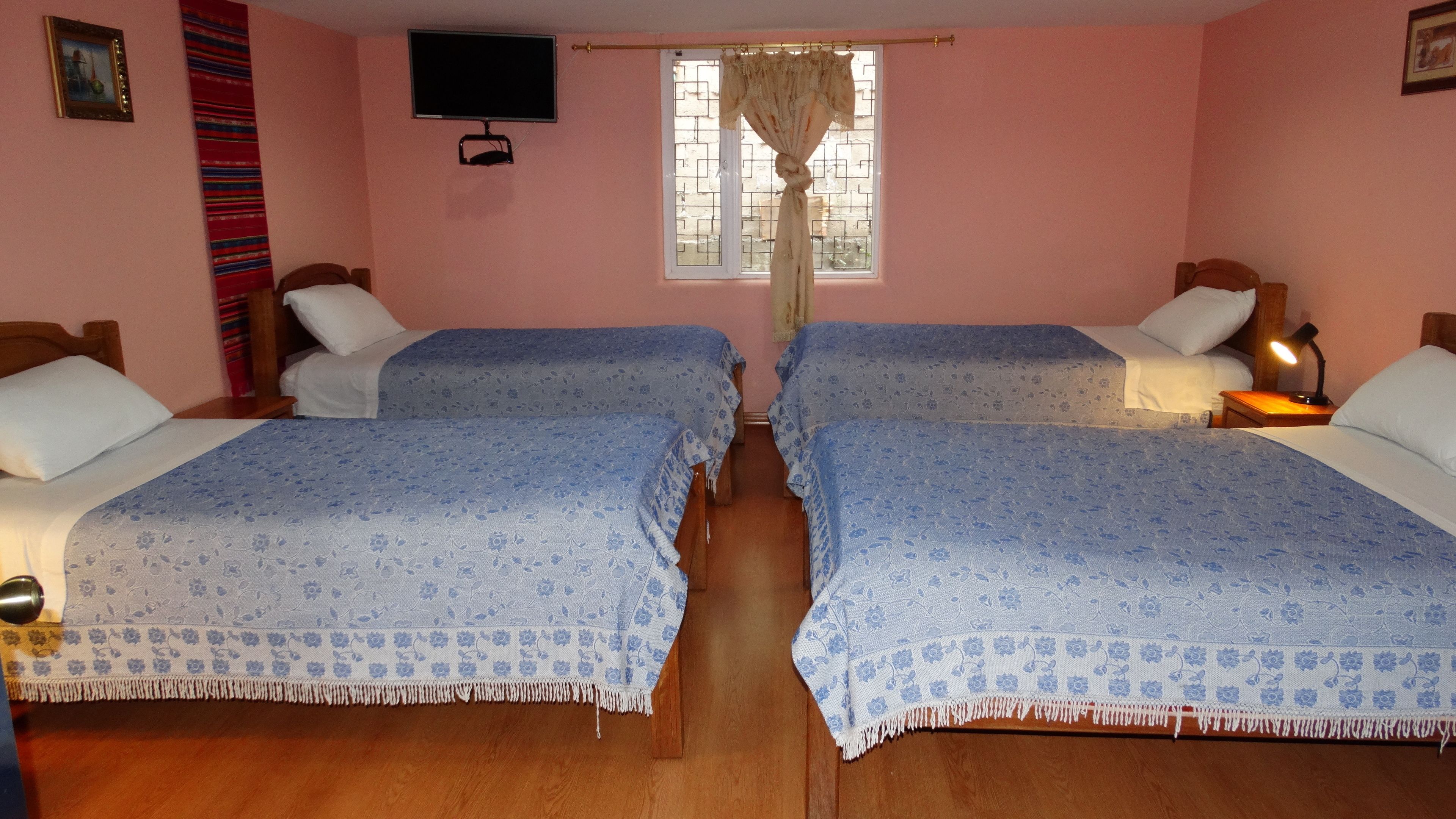 A room with two beds, a television mounted on the wall, a window with curtains, and a wooden floor.