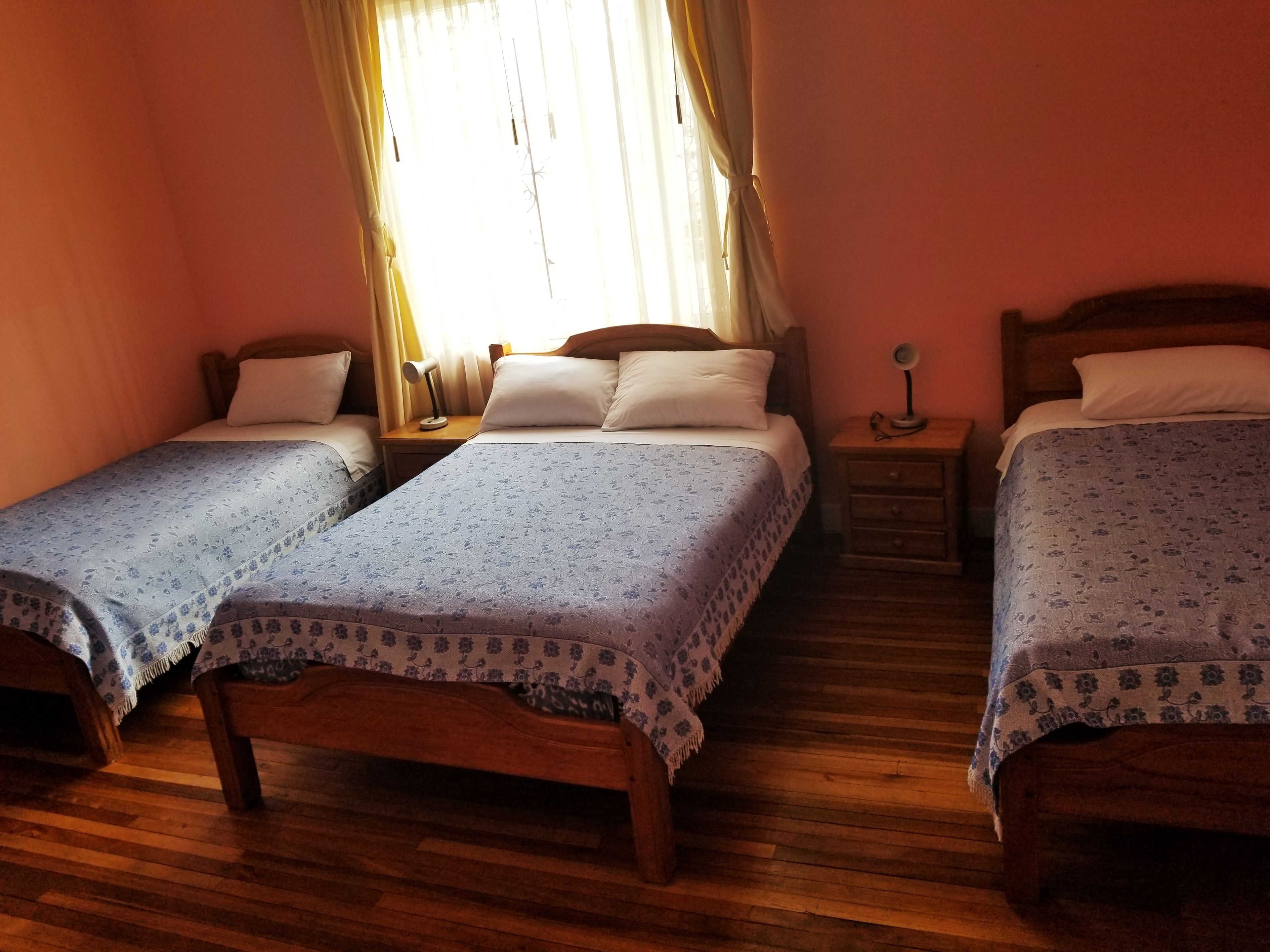 Una habitación con dos camas de madera, cada una con su mesita de noche y una lámpara a juego. El suelo es de madera y hay una ventana con cortinas.