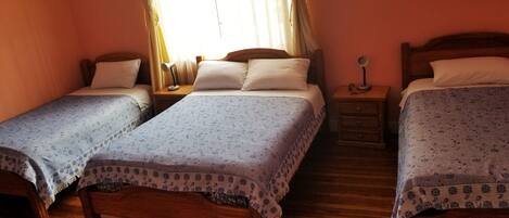 Una habitación con dos camas de madera, cada una con su mesita de noche y una lámpara a juego. El suelo es de madera y hay una ventana con cortinas.