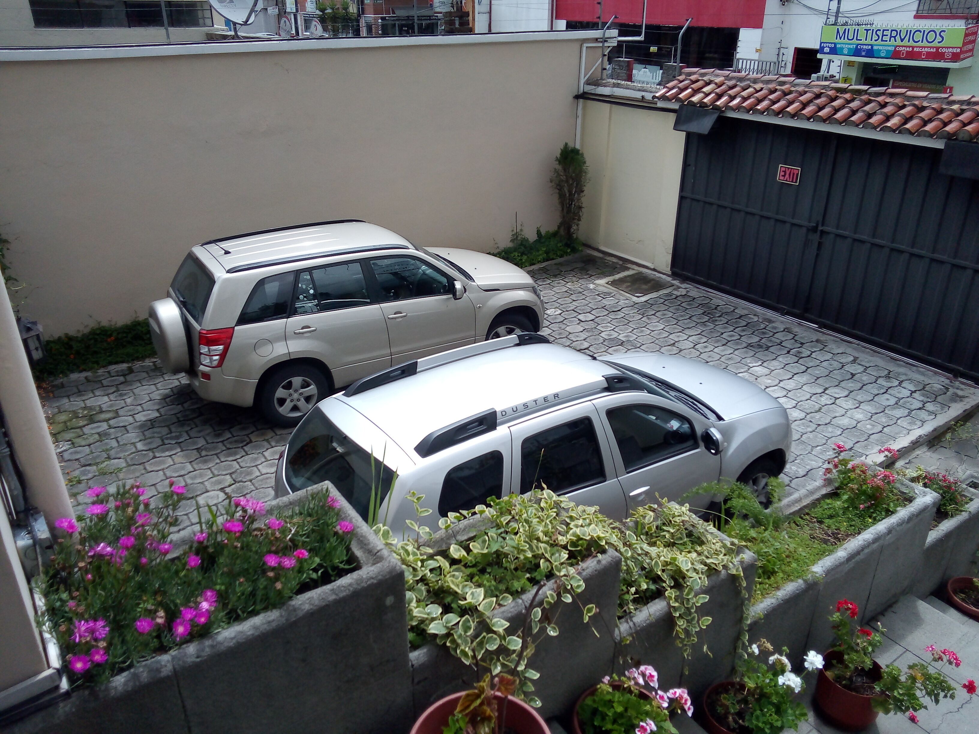 A parking area with two SUVs parked next to a building with a tiled roof and a sign reading "MULTISERVICIOS".
