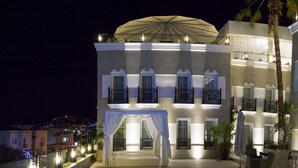 Un área junto a la piscina con un diseño geométrico, un espacio de descanso cubierto con cortinas blancas y un edificio con balcones y una estructura en forma de cúpula.