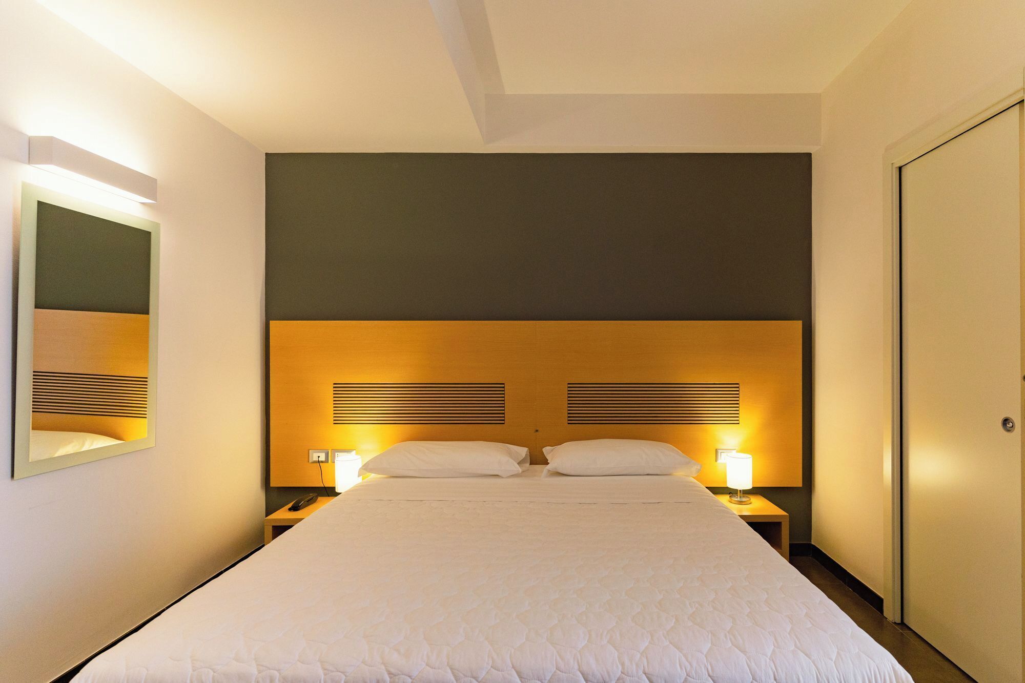 Une chambre d’hôtel avec un grand lit, deux tables de chevet, une commode, un miroir et une porte.