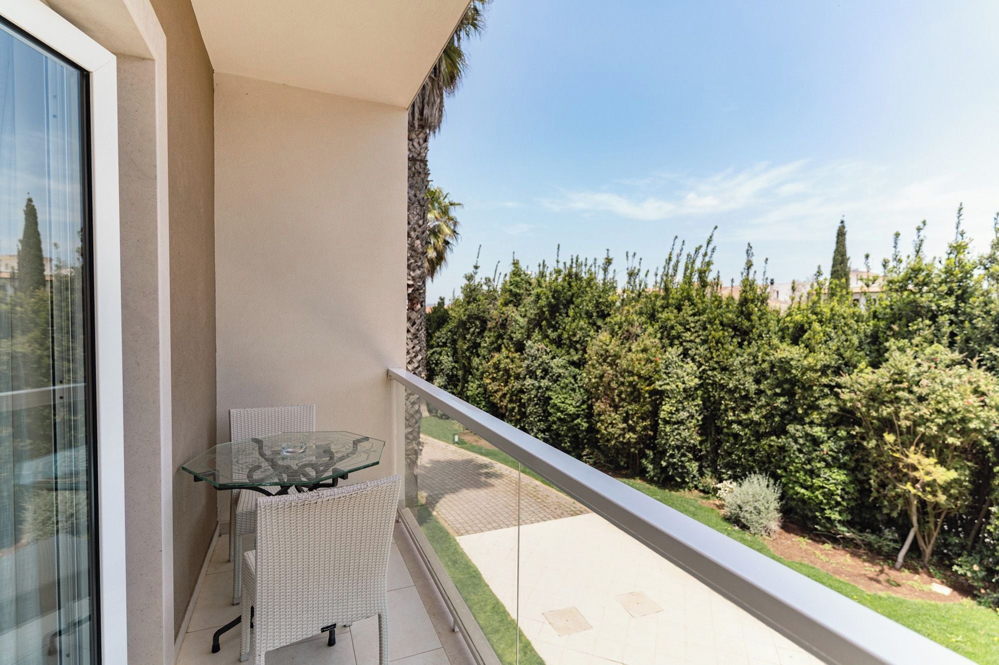 A balcony with a glass table and wicker chairs, overlooking a well-maintained garden and a clear sky.