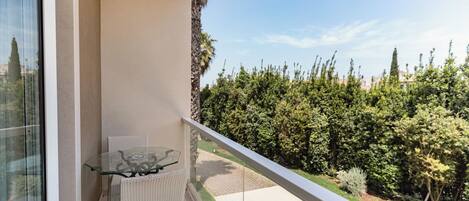 A balcony with a glass table and wicker chairs, overlooking a well-maintained garden and a clear sky.