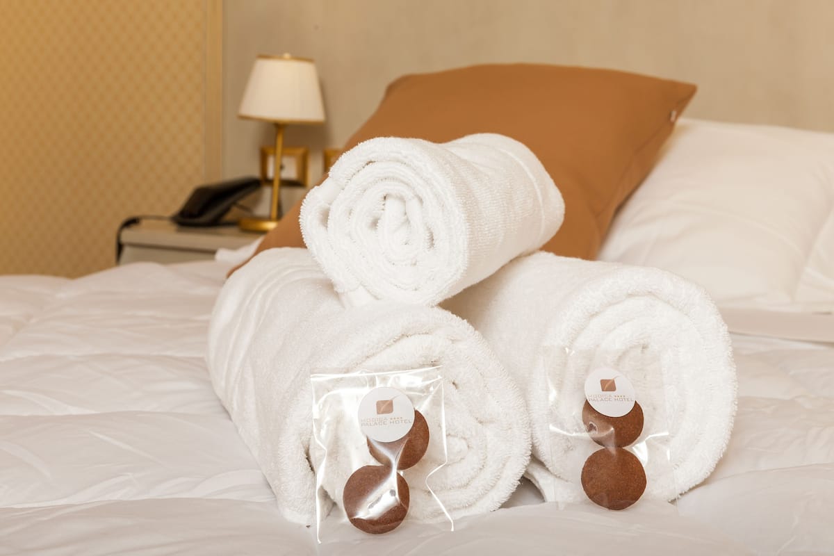 A neatly made bed with rolled white towels and brown slippers, a bedside lamp, and a telephone on a nightstand.