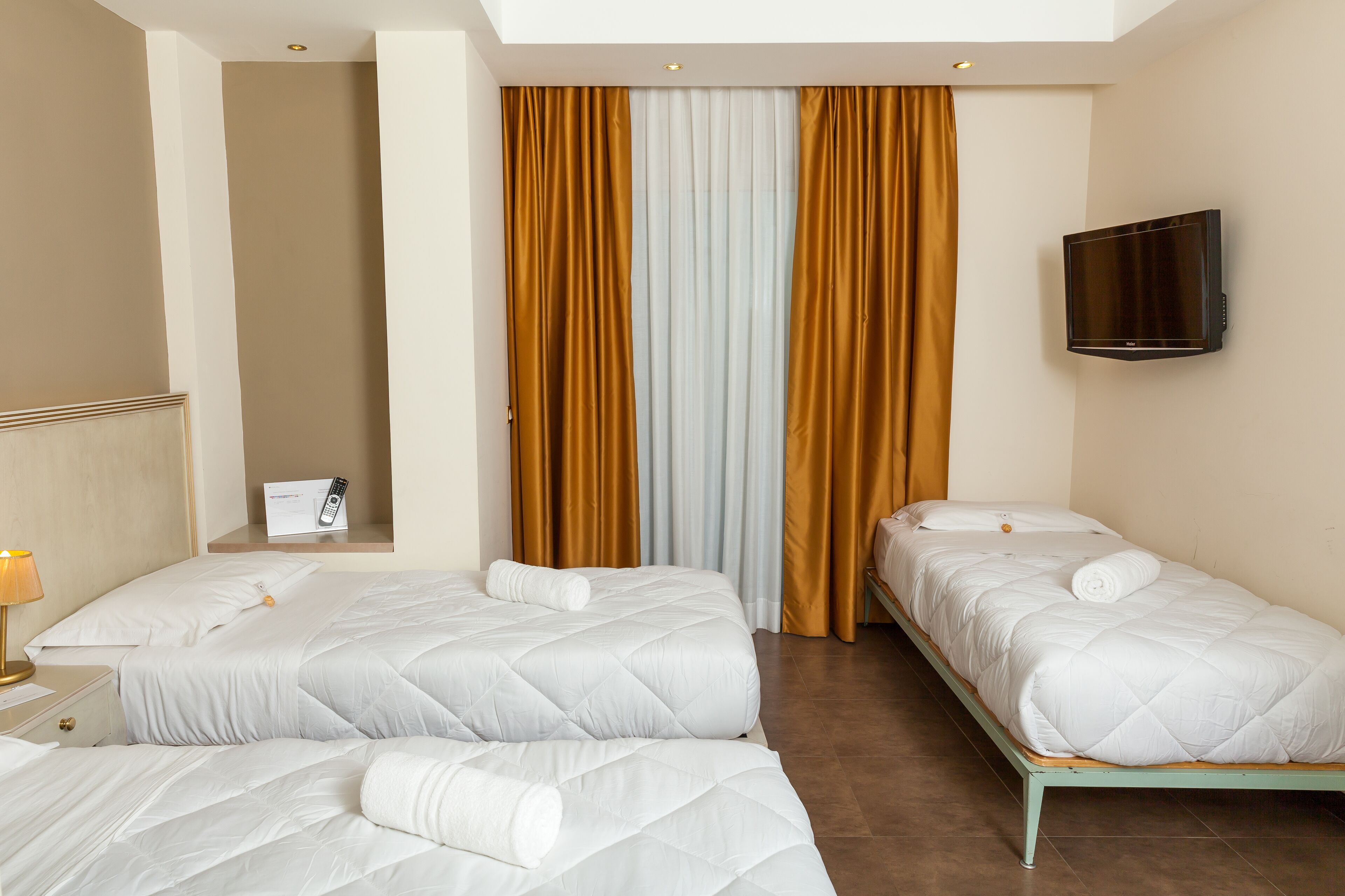 Ein Hotelzimmer mit zwei Betten, einem an der Wand montierten Fernseher und einem Fenster mit Vorhängen.