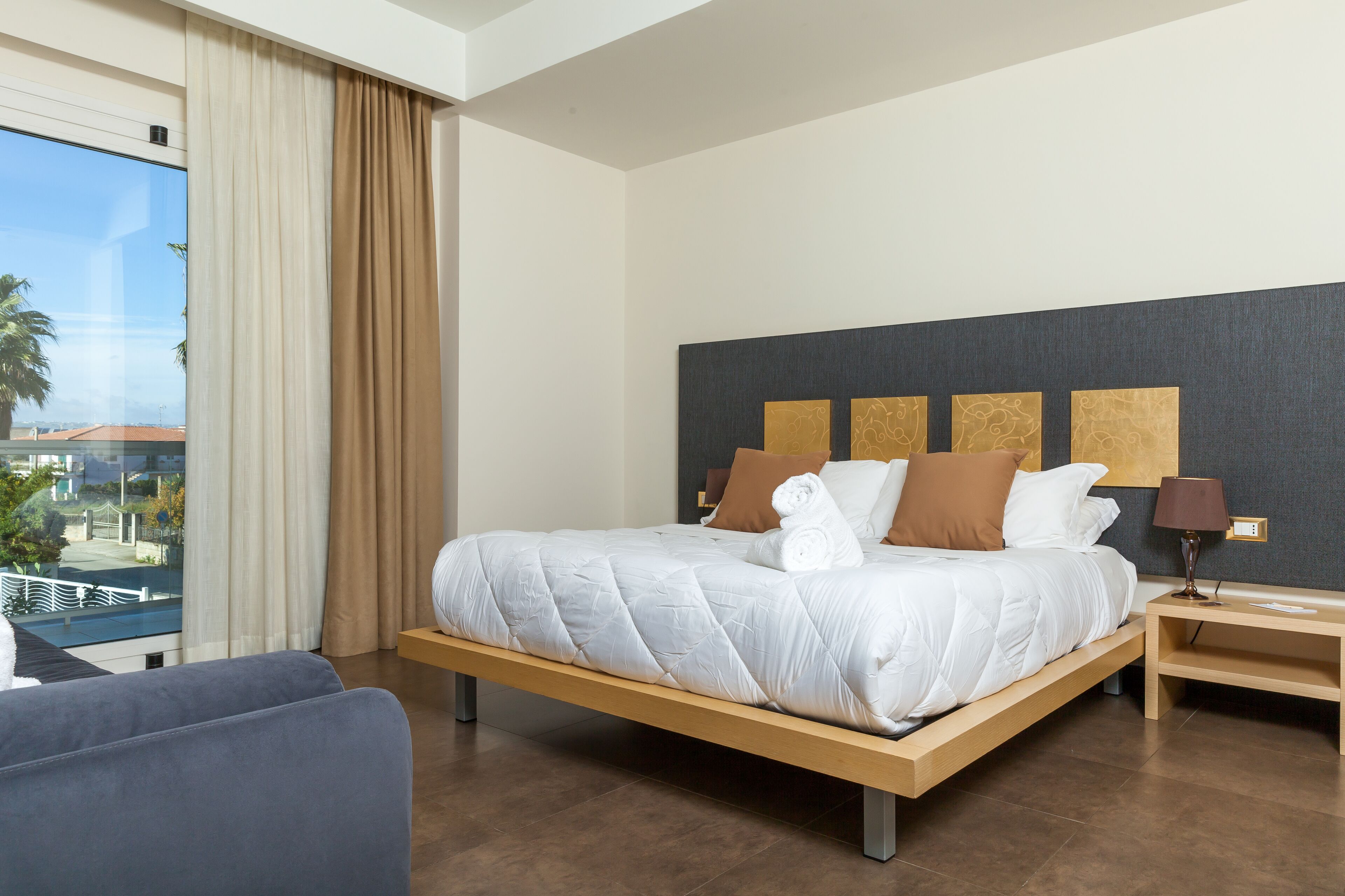 A hotel room with a large bed, a view of the outdoors, a wooden nightstand, and a lamp.