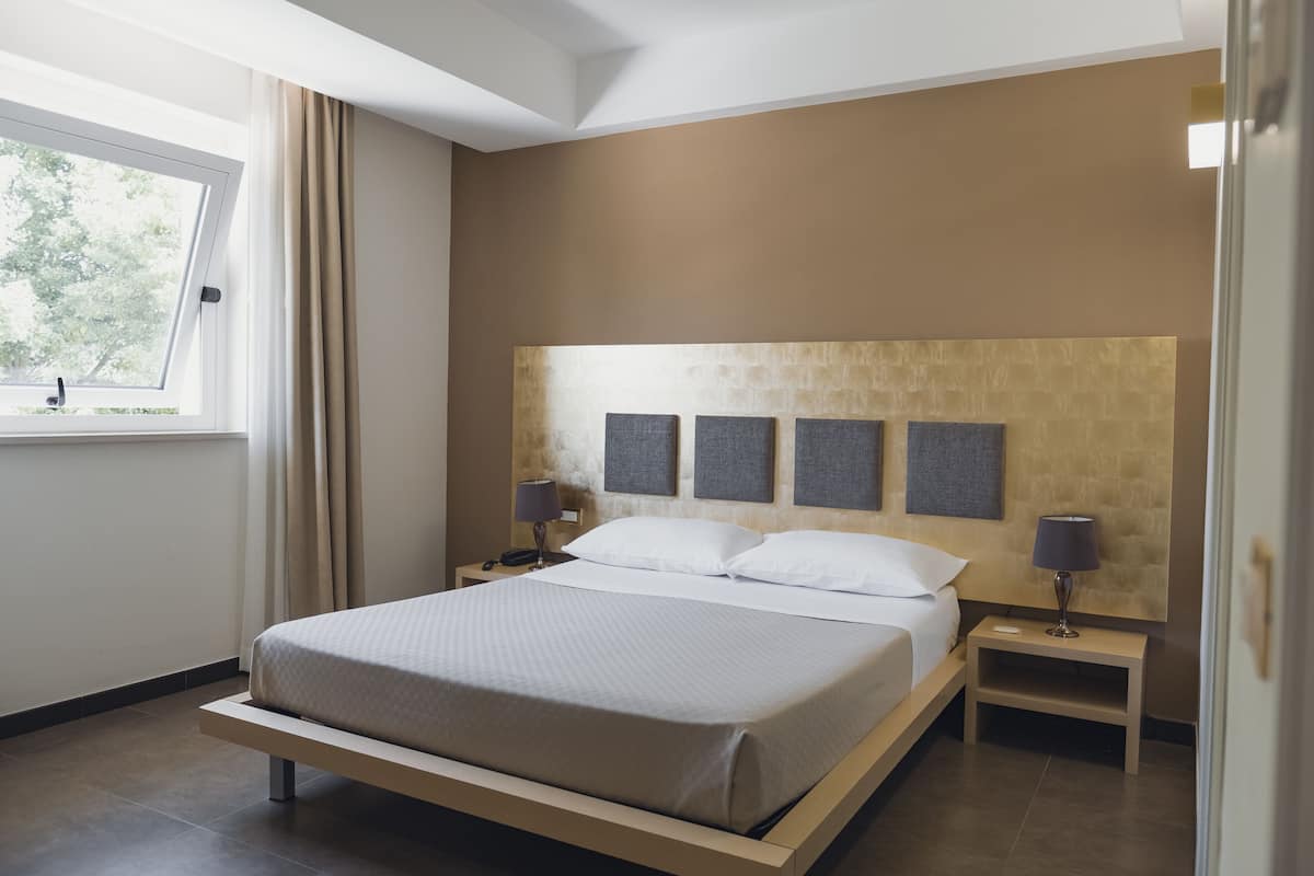 A modern bedroom with a large bed, two bedside tables, a headboard with artwork, and a window with curtains.
