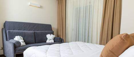 Una habitación de hotel con cama, sofá gris y ventana con cortinas.