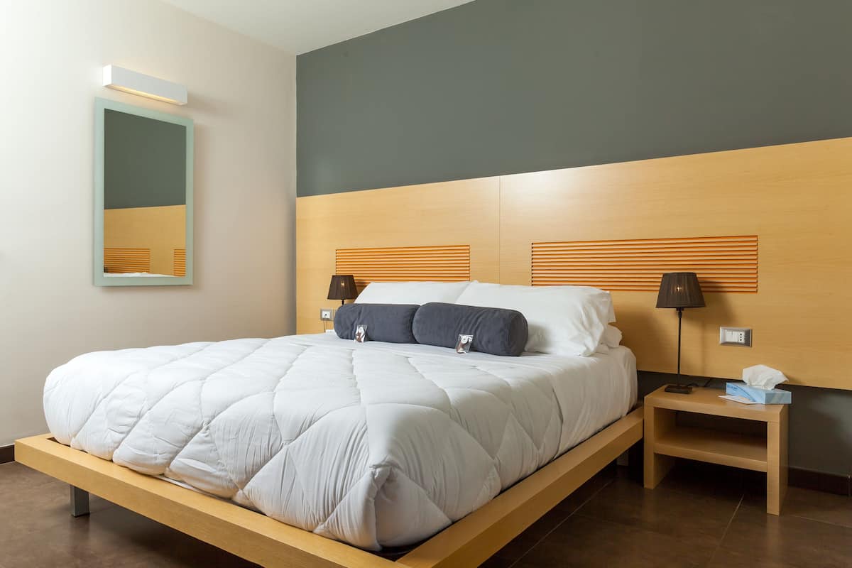 A modern hotel room with a wooden bed frame, a bedside table with a lamp, and a mirror on the wall.